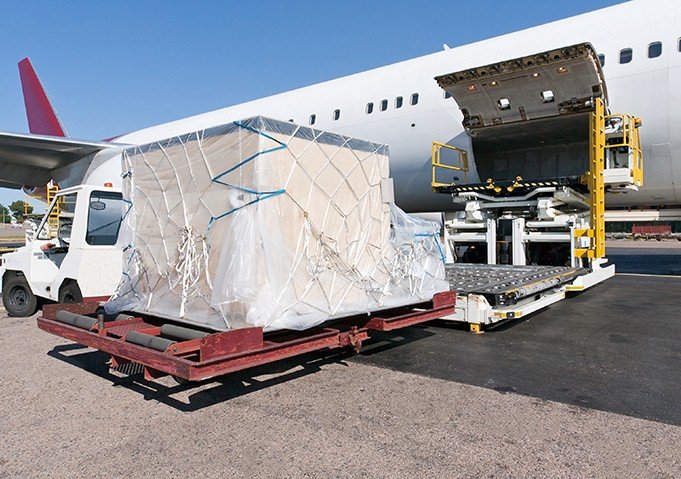 Una gran carga paletizada cubierta con plástico y redes siendo cargada en el compartimiento de carga de un avión de carga en la pista.