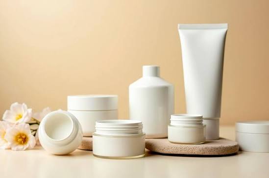 Colección de envases blancos para cosméticos, incluyendo frascos, tubos y un frasco abierto, junto a flores blancas sobre una superficie clara.